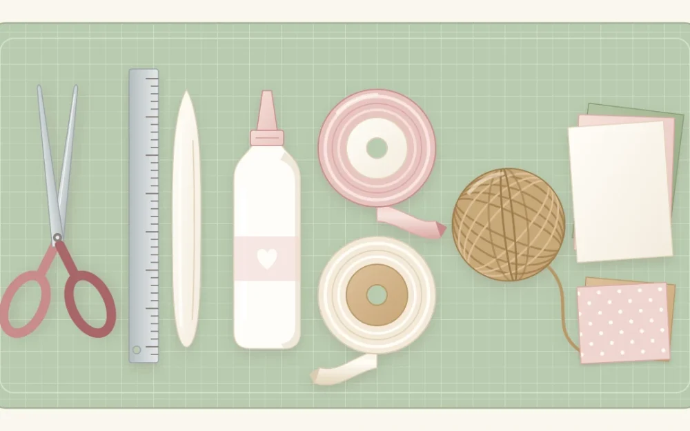 Scissors, a metal ruler, a bone folder, a glue bottle, blush and ivory ribbon spools, twine and cardstock in a row on a sage cutting mat